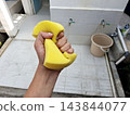 Close up of hand squeezing wet yellow sponge for cleaning activity near outdoor area with ceramic tiles water taps and household equipment. 143844077