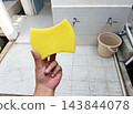 Human hand grasping clean yellow sponge for washing tasks near outdoor tiled area with water taps and utility bucket for cleaning purposes. 143844078