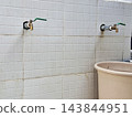 Two stainless steel ball valve taps extending from light gray ceramic tiled wall above beige plastic basin in utility area or public washroom space. 143844951