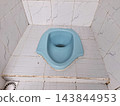 Simple light blue squat toilet in a small tiled bathroom, featuring white floor and wall tiles with a bottom drain. 143844953
