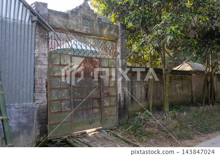 Old rusty iron entrance gate and concrete arch of a rural home. Traditional village house boundary wall surrounded by green trees. Old rusty iron entrance gate and concrete arch of a rural home. Traditional village house boundary wall surrounded by green trees. 143847024