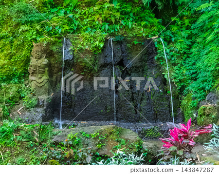 Sacred water spring with stone Hindu guardian statue and water jets on dark volcanic rock wall in tropical jungle, Bali, Indonesia 143847287
