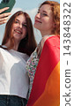 Vertical video. Young lesbian couple taking selfie in the city. Two happy girlfriends pose with rainbow flag. Lgbt woman, showing love to each other. 143848322
