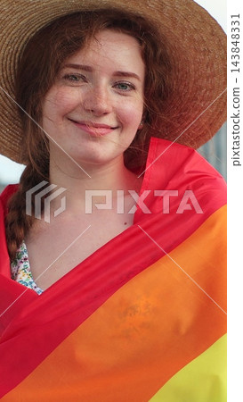 Vertical video. Portrait of a young woman in a hat with long red hair covering herself in lgbt flag while looking to camera. Vertical video. Portrait of a young woman in a hat with long red hair covering herself in lgbt flag while looking to camera. 143848331
