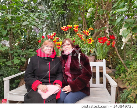 Two older women sit together on a white bench among flowering plants, enjoying companionship during a visit to a lush greenhouse 143849723