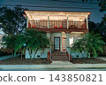 White Christmas lights illuminate the upper balcony and roof of a two-story house. Palm trees, red chairs, and a hammock create a tropical holiday scene. 143850821