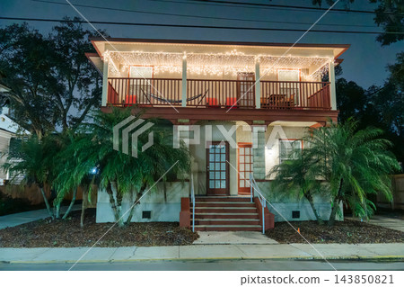 White Christmas lights illuminate the upper balcony and roof of a two-story house. Palm trees, red chairs, and a hammock create a tropical holiday scene. 143850821