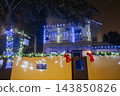 Blue and white Christmas lights illuminate a two-story house, balcony, fence, and large tree. Red bows and green garland decorate the entrance on a festive Florida evening. 143850826