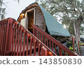 A weathered wooden boat rests on display beneath a curved green protective canopy. Red stairs, string lights, and moss-covered trees surround the historic vessel. 143850830