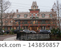 A symmetrical Victorian hotel displays wreaths, bows, lights, and colorful Christmas decorations. Bare trees and parked vehicles frame the historic building on an overcast day. 143850857