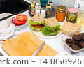 Homemade burgers are assembled on a kitchen counter with buns, lettuce, tomato, onion, cooked patties, and jars of condiments during meal preparation 143850926