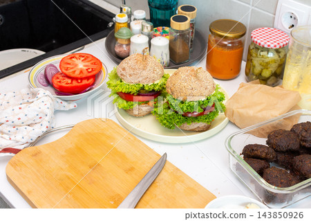 Homemade burgers are assembled on a kitchen counter with buns, lettuce, tomato, onion, cooked patties, and jars of condiments during meal preparation Homemade burgers are assembled on a kitchen counter with buns, lettuce, tomato, onion, cooked patties, and jars of condiments during meal preparation 143850926