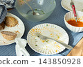 Used plates, bowls, a fork, and a spoon sit on a striped tablecloth after a meal, with bread and sauce left on dishes 143850928