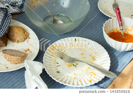 Used plates, bowls, a fork, and a spoon sit on a striped tablecloth after a meal, with bread and sauce left on dishes Used plates, bowls, a fork, and a spoon sit on a striped tablecloth after a meal, with bread and sauce left on dishes 143850928