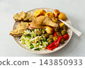 A plate holds baked chicken leg, potatoes, fresh salad, roasted Brussels sprouts, and chickpea bread. This meal is ready to eat at a dining table 143850933
