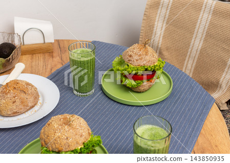 At breakfast, homemade burgers with lettuce and tomato sit on green plates on a wooden table, with green drinks beside them in a dining area 143850935