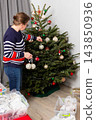 A person is standing next to a Christmas tree. They hold two decoration balls in their hands. The tree is decorated with lights and ornaments 143850936