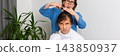 A woman is cutting her husband's hair at home. They are in their living room with plants around. It is a casual afternoon, and he sits in a chair covered with a cape, banner 143850937