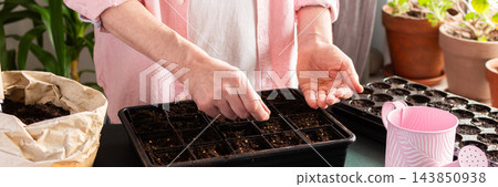 A man sows tomato seeds into a seed tray at home, surrounded by gardening tools and seed packets. He is engaged in spring garden preparation, nurturing plants from seed stage, banner 143850938