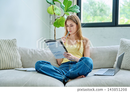 Young student girl study online, sitting at home on couch with laptop, textbook Young student girl study online, sitting at home on couch with laptop, textbook 143851208