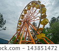 A large orange Ferris wheel rises above trees at an amusement park. Bright yellow gondolas stand against a dramatic cloudy sky. 143851472