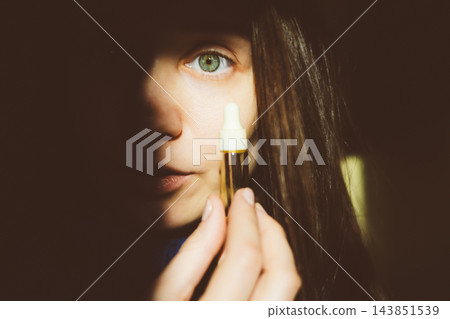 Portrait of a young woman with natural skin holding a glass pipette with a serum drop. Authentic beauty and skincare routine. Wellness aesthetic. 143851539