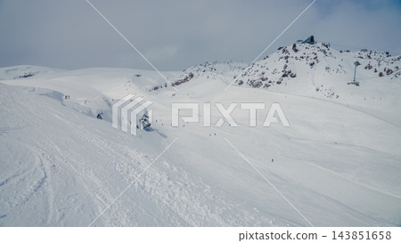 Snow-covered mountain slope with skiers near rocky peak under overcast sky, Caucasus Elbrus region Snow-covered mountain slope with skiers near rocky peak under overcast sky, Caucasus Elbrus region 143851658