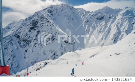 Skiers descending sunlit snowy slope below rugged Mount Elbrus peaks in Caucasus panorama Skiers descending sunlit snowy slope below rugged Mount Elbrus peaks in Caucasus panorama 143851684