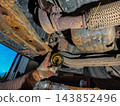 Mechanic in work gloves replacing a dirty oil filter with old oil draining, revealing rusty chassis components and exhaust system of a vehicle lifted for routine service. 143852496