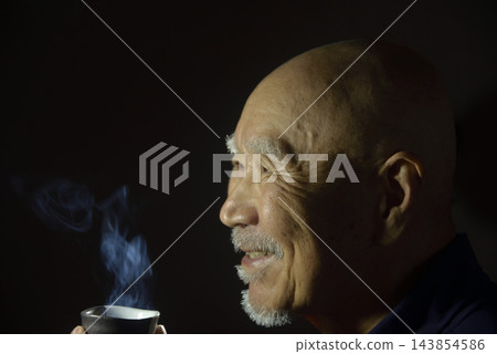 一位老者面帶微笑地喝著杯中熱氣騰騰的清酒。 一位老者面帶微笑地喝著杯中熱氣騰騰的清酒。 143854586