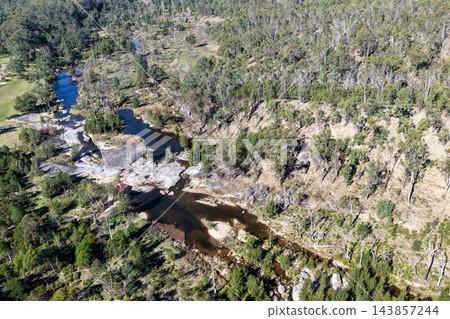 Aerial photograph of a shallow river flowing over broad rock ledges into pools, surrounded by native woodland and rugged bushland. 143857244