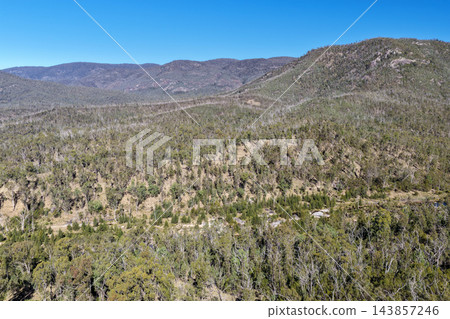 Aerial photograph of rugged forested hills and rocky ridgelines rising above dry native bushland beneath a clear blue sky. Aerial photograph of rugged forested hills and rocky ridgelines rising above dry native bushland beneath a clear blue sky. 143857246