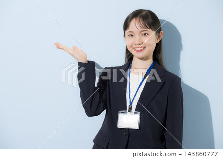 [淺藍色背景] 一位二十出頭的年輕日本女子，身穿西裝，脖子上掛著員工證，正在這裡指引人們。 143860777