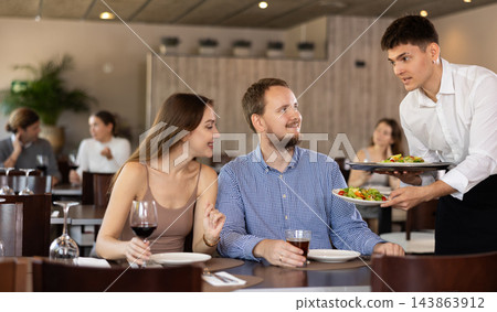 Friendly smiling waiter bringing dishes to guests of restaurant Friendly smiling waiter bringing dishes to guests of restaurant 143863912