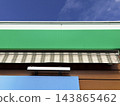 Low-angle view of a storefront with a green panel and a striped awning against a blue sky. The building facade 143865462