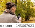 Rear view of a man with a hair bun in a beige coat holding a smartphone while standing on a park path under yellow autumn trees. Concept of a mindful walk and digital life balance. 143867023