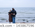 Middle aged couple hugging and looking at the Baltic Sea from behind, standing on a pebble beach in warm coats on a gloomy autumn day. Concept of support between spouses facing a big life change. 143867596