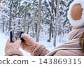 Teenage girl in fuzzy earmuffs and mittens holding a smartphone with her own selfie on the screen in a snowy forest. Concept of social media, teen selfie culture and winter trip memories. 143869315