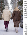 Woman and teenage girl holding hands and walking away along a snowy forest road past a parked car. Concept of winter road trip, family bonding and travel by car through nature. 143869316