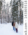Man and two young people walking one behind another along a snowy forest trail in warm winter clothes. Concept of family hiking, winter vacation and shared adventure outdoors. 143869318