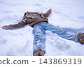 Teenage girl lying in fresh snow making a snow angel with arms outstretched in a brown jacket, scarf and jeans. Concept of carefree winter fun, childhood joy and active family vacation. 143869319