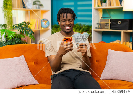 African American young man on home sofa counting cash and checking amount on smartphone with smile 143869459