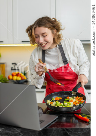 Young woman cooking veggie stir fry while watching laptop recipe video for diet meal in home kitchen 143869461