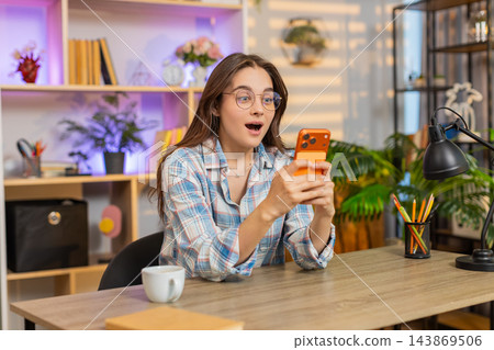 Young woman on home table reading smartphone good news feeling surprise joy luck and success indoors Young woman on home table reading smartphone good news feeling surprise joy luck and success indoors 143869506