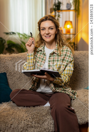 Young woman on home sofa writing personal goals in notebook smiling with inspiration and check mark 143869528