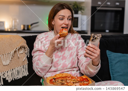 Adult woman enjoys pizza slice while texting on smartphone at night smiles cozy mood friendly chat Adult woman enjoys pizza slice while texting on smartphone at night smiles cozy mood friendly chat 143869547