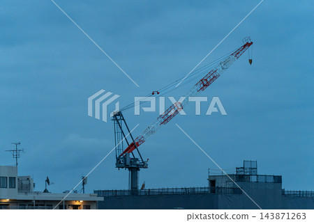 在建築工地上，一架巨大的紅白相間的起重機高聳入雲，背景是泛著藍色的暮色天空。 143871263