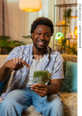 African American young man on home sofa holds cash plans purchase and celebrates online casino win 143872910