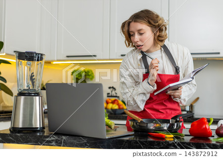 Young woman cooking healthy diet meal while checking laptop recipe and writing notes in home kitchen Young woman cooking healthy diet meal while checking laptop recipe and writing notes in home kitchen 143872912