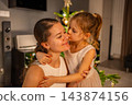 A girl kisses a woman on the cheek in front of a decorated Christmas tree. A holiday embrace expresses affection during New Year celebrations at home. 143874156